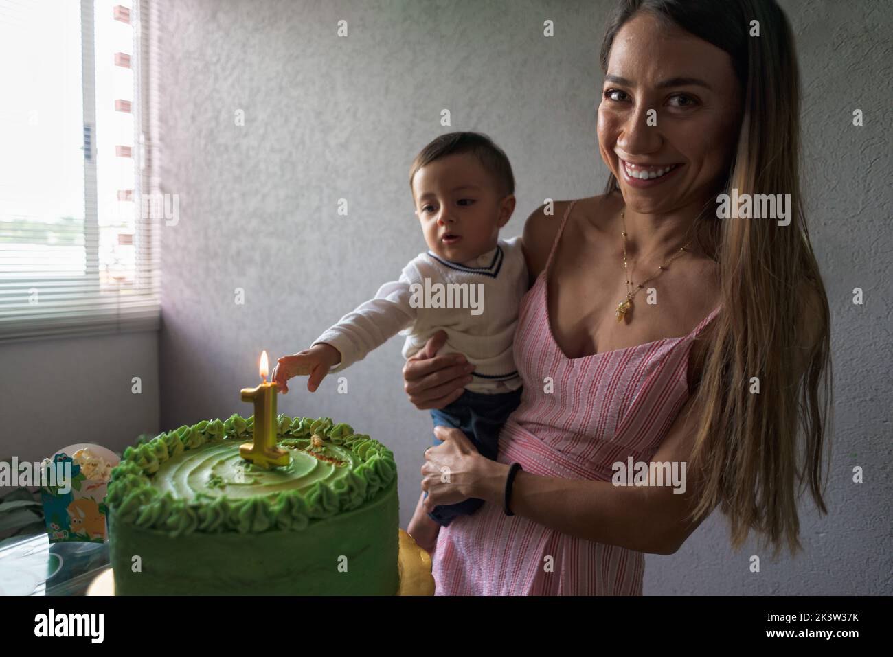 Encantado madre étnica mirando la cámara sosteniendo bebé niño mientras celebraba el cumpleaños con pastel de fiesta decorado con velas encendidas Foto de stock