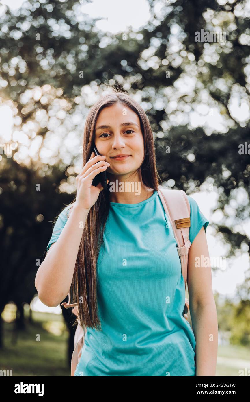 Adolescente niña estudiante con camisa de aguamarina, haciendo una llamada telefónica a su novio en el parque. Retroiluminación solar Foto de stock