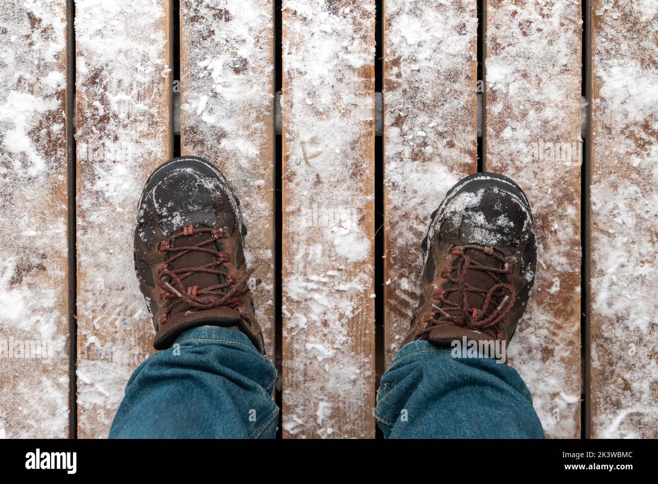 Foto de cerca de los pies masculinos en botas de trekking parados sobre suelo de madera nevado, vista en primera persona Foto de stock
