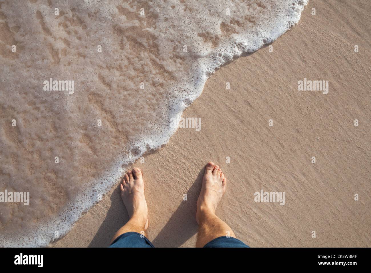 Piernas descalzas de un hombre parado en una playa en la arena costera húmeda en un día soleado, vista superior Foto de stock