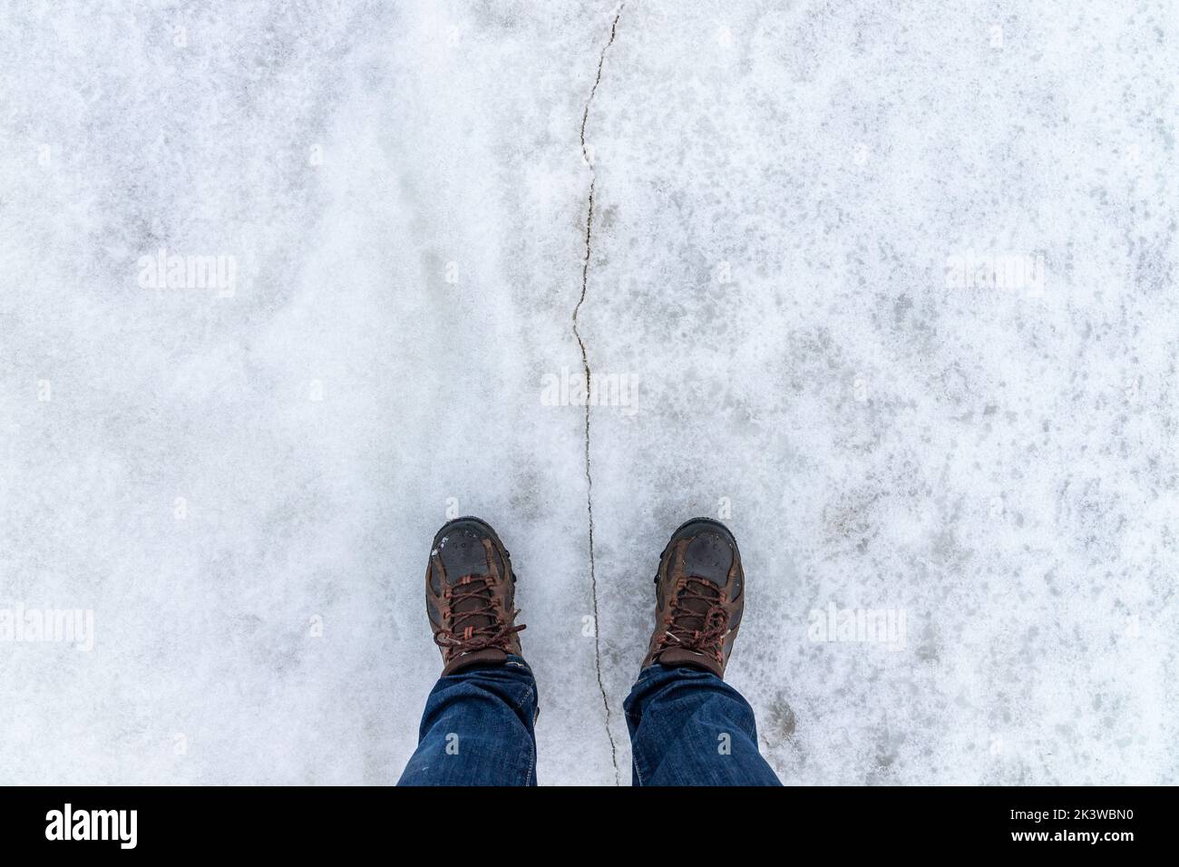 Los pies masculinos están de pie sobre un hielo nevado con grietas, vista en primera persona, vista superior Foto de stock