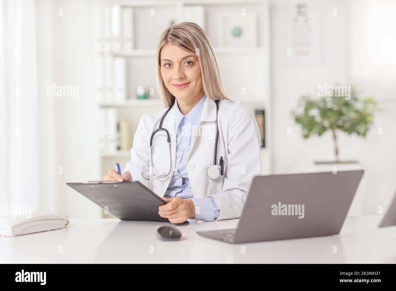 Joven doctora sentada en un consultorio y escribiendo un documento en un escritorio con ordenador portátil Foto de stock
