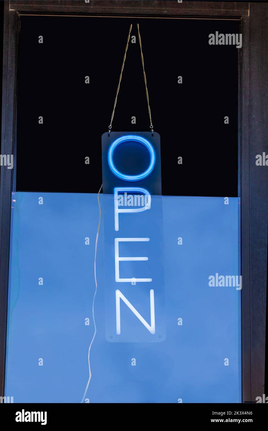 Letrero de neón ABIERTO que cuelga de la puerta hacia los negocios con el cielo azul reflejado en la puerta de cristal y el cable eléctrico colgando del letrero Foto de stock