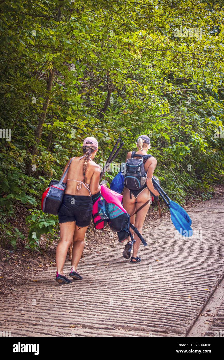 Kayakers sexy - dos niñas en la rampa de barco caminar con la vida chalecos oars backpacks y otro equipo de kayak - uno tiene en un thong Foto de stock