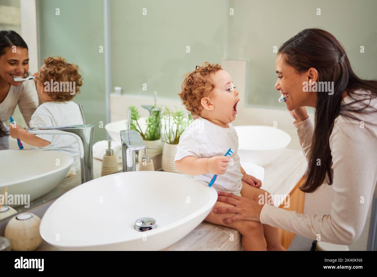 Mamá que enseña a bebé a cepillarse los dientes, en el mostrador del cuarto de baño en casa y una sonrisa limpia en su cara. Una higiene bucal saludable para el niño significa usar al niño Foto de stock