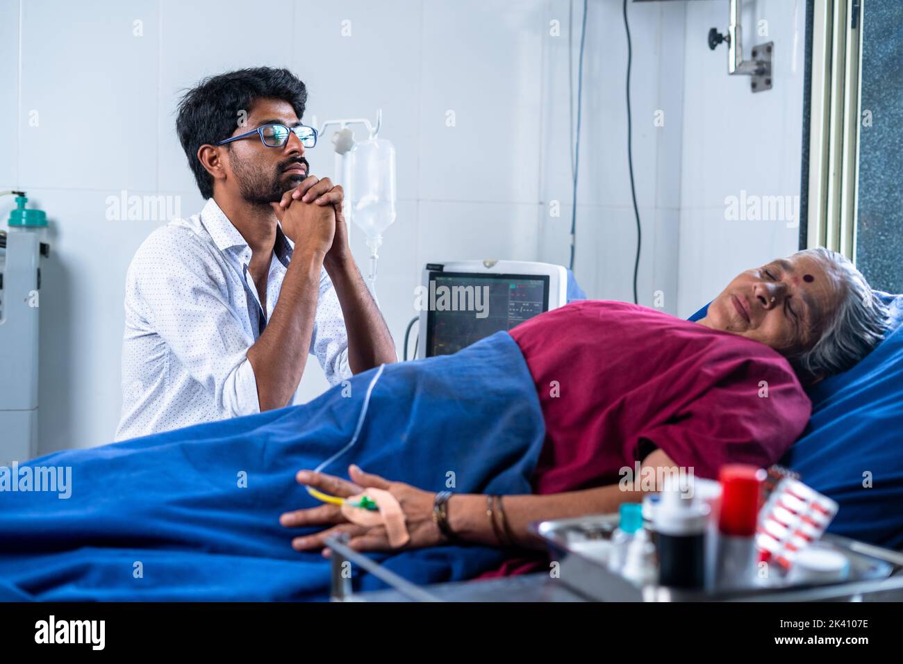 Preocupado hijo indio rezando a dios por la madre enferma para acelerar la recuperación de la salud en el hospital - concepto de atención médica, vínculo incondicional y tratamiento. Foto de stock