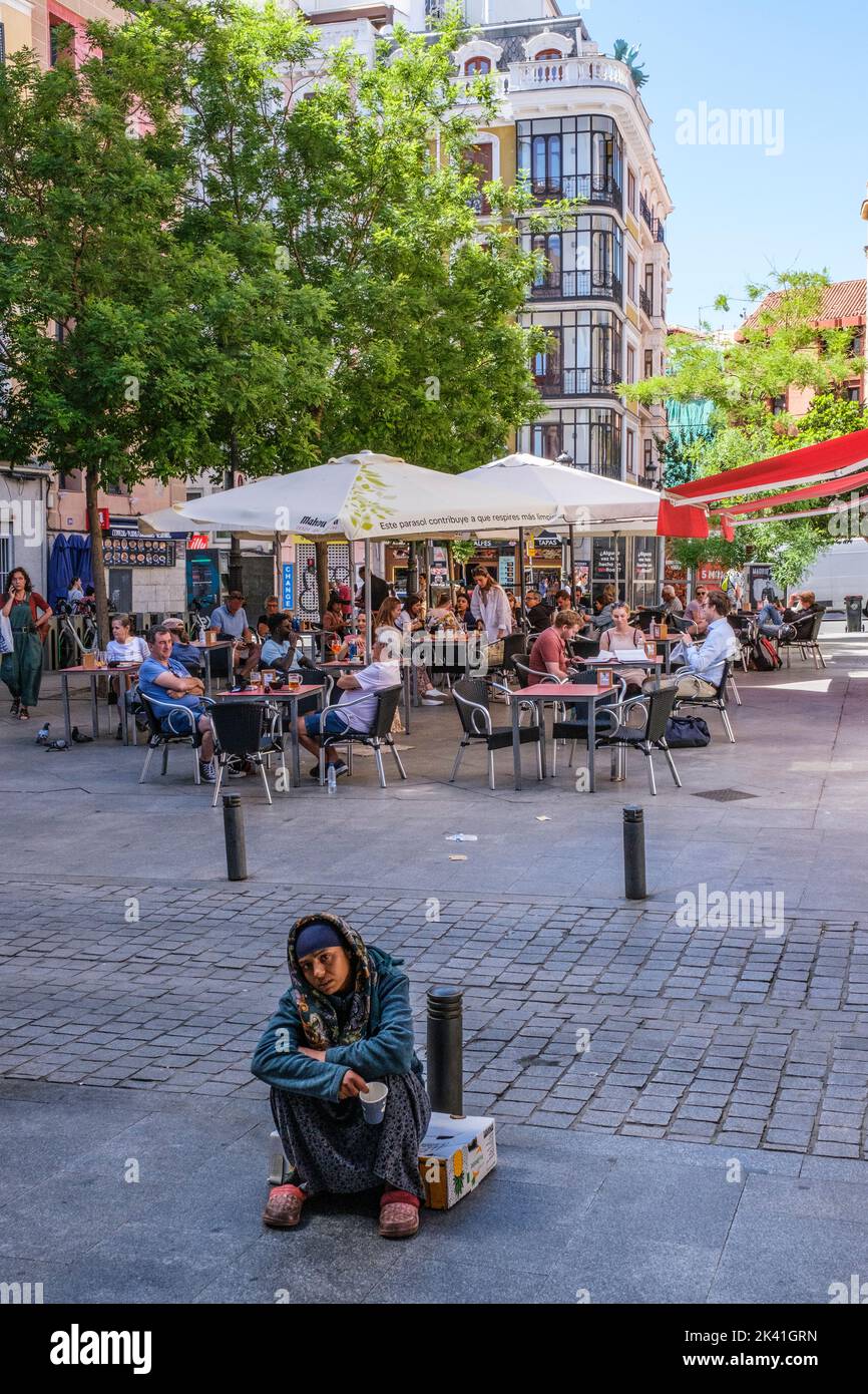 España, Madrid. Mercado de San Miguel. El mendigo espera fuera del mercado mientras los clientes disfrutan del almuerzo en las mesas exteriores del comedor. Foto de stock