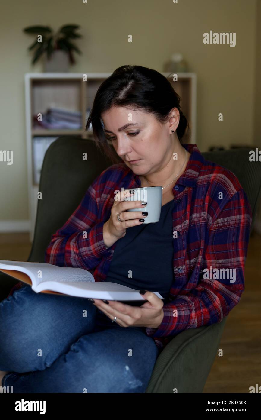Mujer morena Relájate con un libro y una taza de té en el sillón de la casa. Actividad de lectura para adultos hermosas mujeres, disfrutando de un estilo de vida tranquilo interior Foto de stock