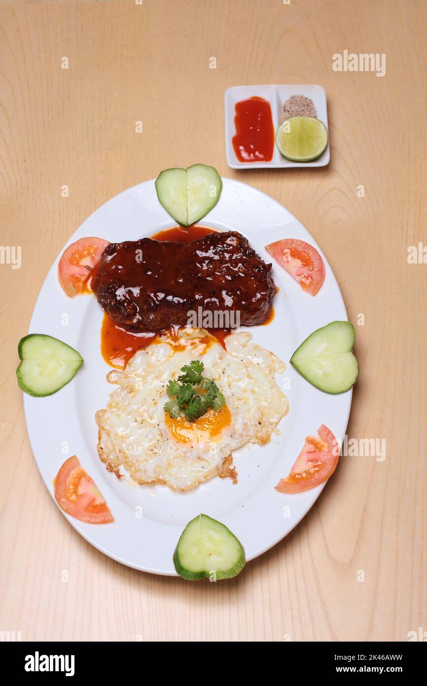 Bistec con huevo frito y verduras se colocan sobre un plato blanco, sobre una mesa de madera, servido con salsa de chile, sal y limón Foto de stock