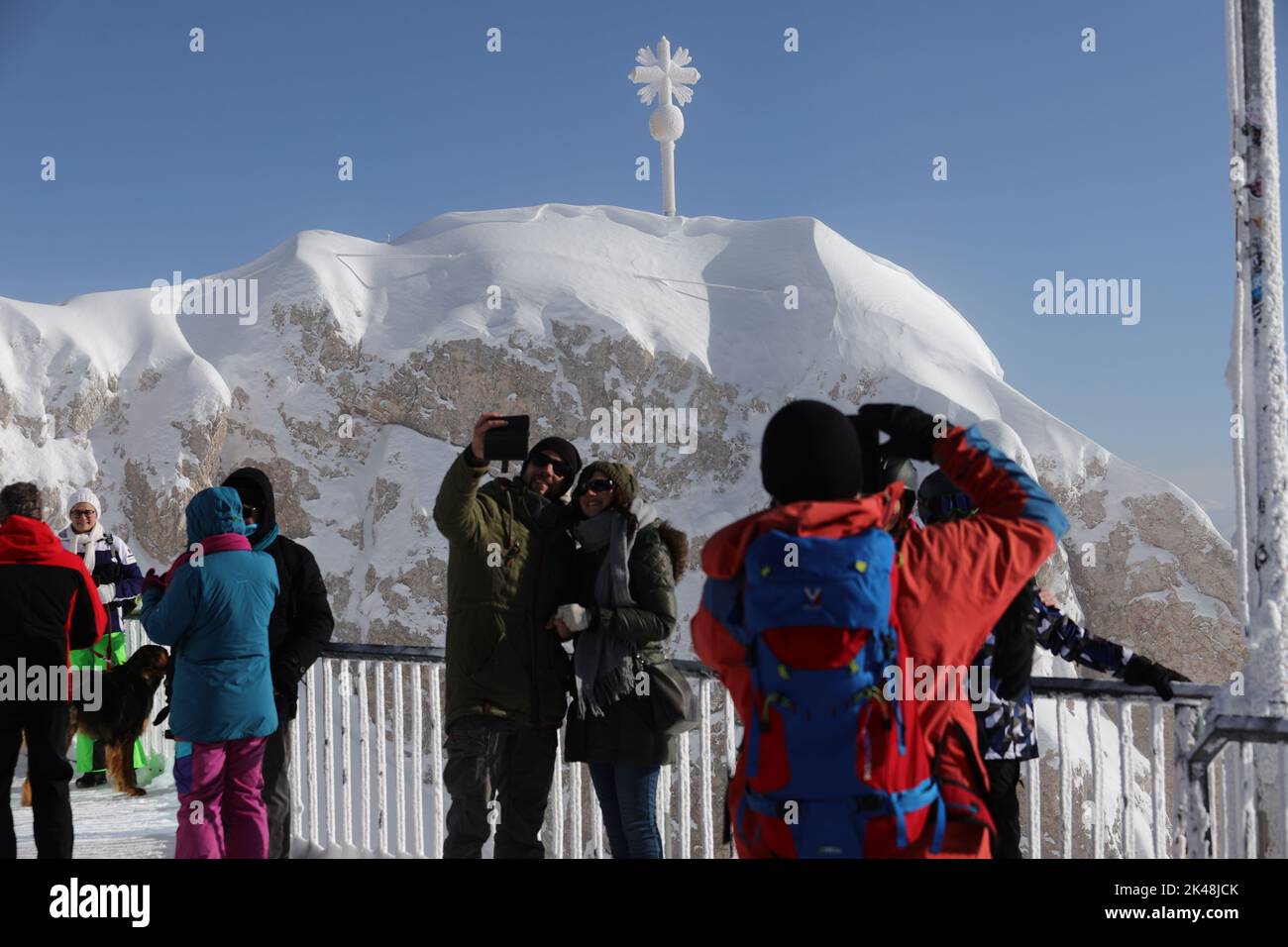Besucher auf der Zugspitze fotografieren das Gipfelkreuz und machen selfies. Es herrscht bittere Kälte, und alle Touristen sind warm angezogen. Das Gipfelkreuz ist mit Raureif überzogen und hebt sich hell gegen den wolkenlosen, blauen Himmel ab Foto de stock