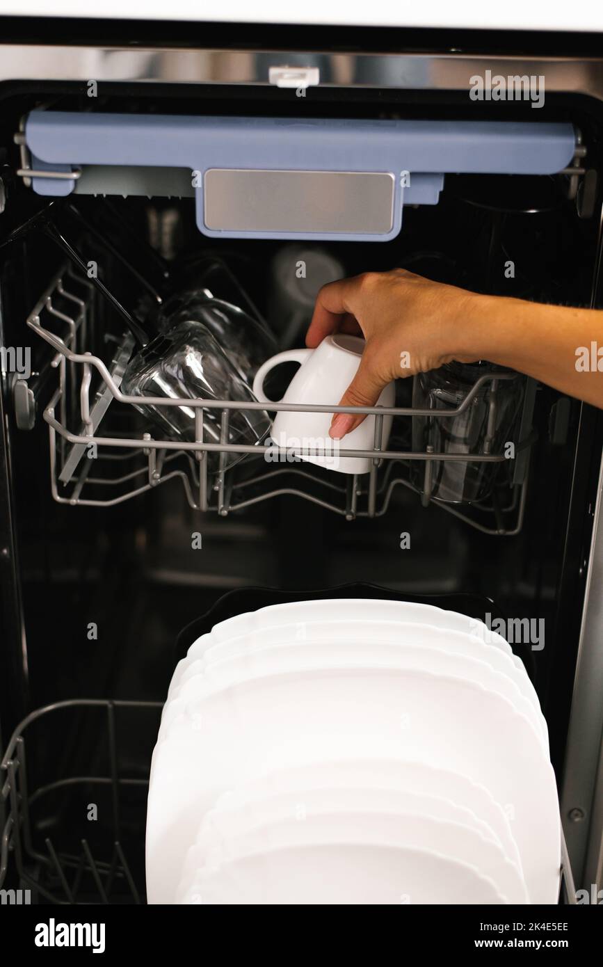 Primer plano de la mano de una mujer cargando platos, vaciándolos o descargándolos de un lavavajillas integrado automático abierto con platos en la cocina. Foto de stock