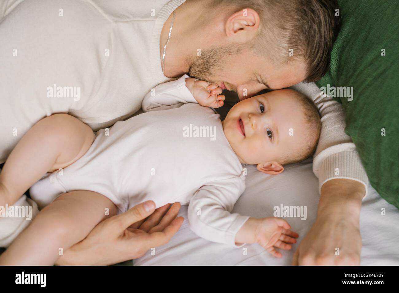 Un padre joven guapo miente en la cama en el dormitorio y abraza a su amante niño de seis meses riendo. El concepto de amor incondicional y paternidad Foto de stock