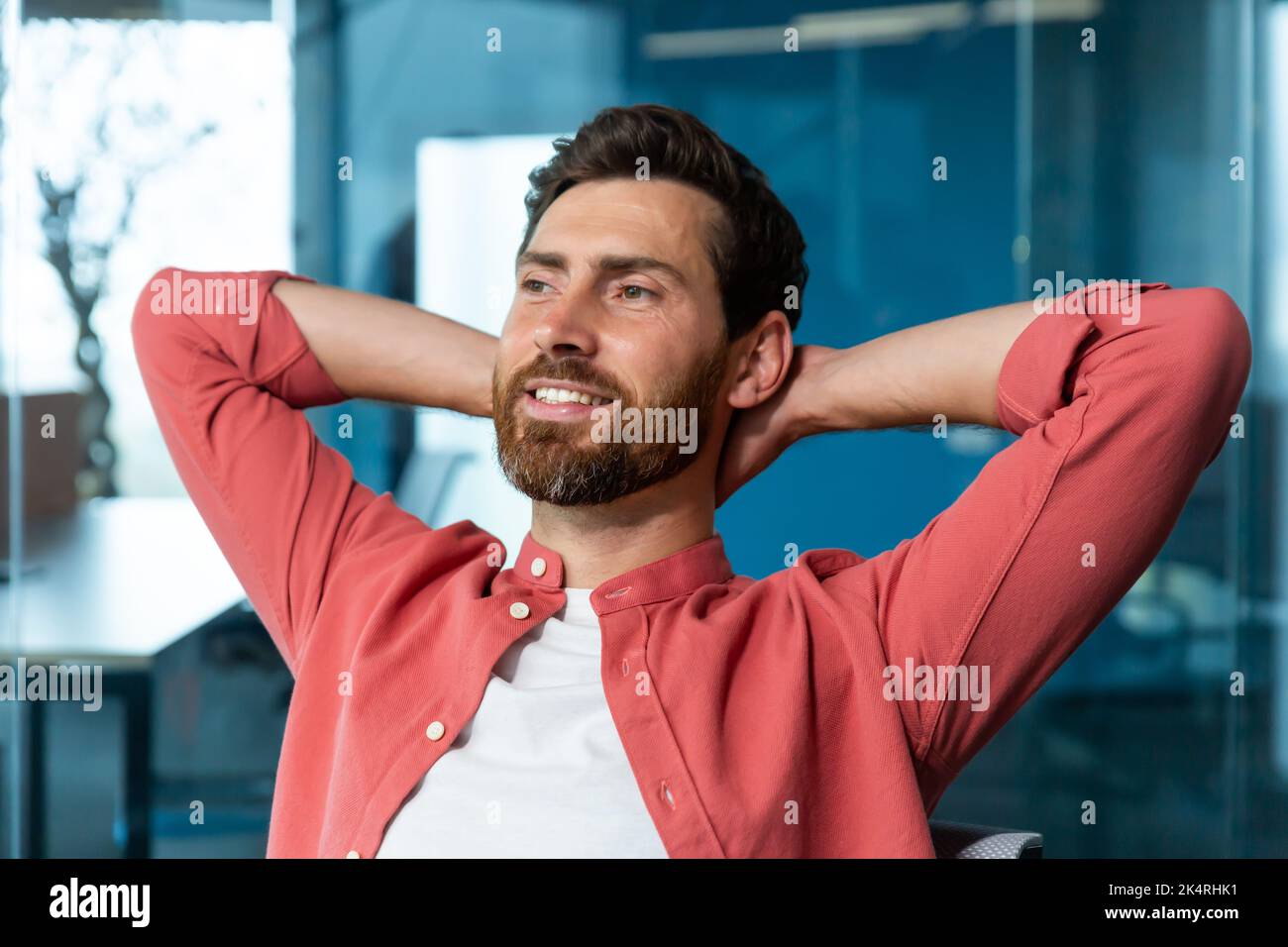 El joven exitoso hombre de negocios startuper descansa en la oficina, el hombre con camisa roja lanza las manos detrás de la cabeza y los sueños visualiza, el diseñador piensa en un nuevo proyecto. Foto de stock