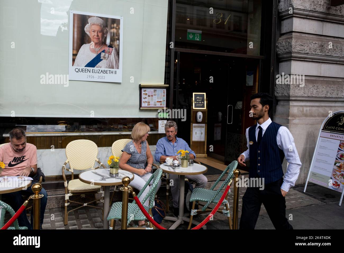 Imagen de la Reina en la ventana del Cafe Concerto mientras los clientes disfrutan de un café en el exterior tras la muerte de la Reina Isabel II, dejada como señal de respeto el 12th de septiembre de 2022 en Londres, Reino Unido. El rostro de la ahora difunta Reina será una imagen permanente durante los siglos venideros. Foto de stock
