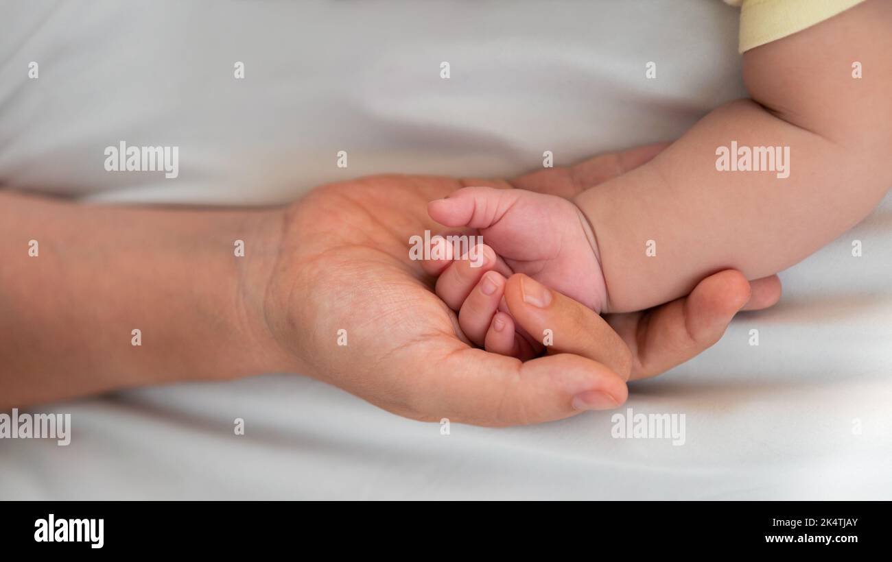 Madre joven sosteniendo la mano del bebé y mostrando el dedo y la uña en la sábana blanca. Primer plano del bebé durmiendo en la habitación de la madre. Familia y casa co Foto de stock