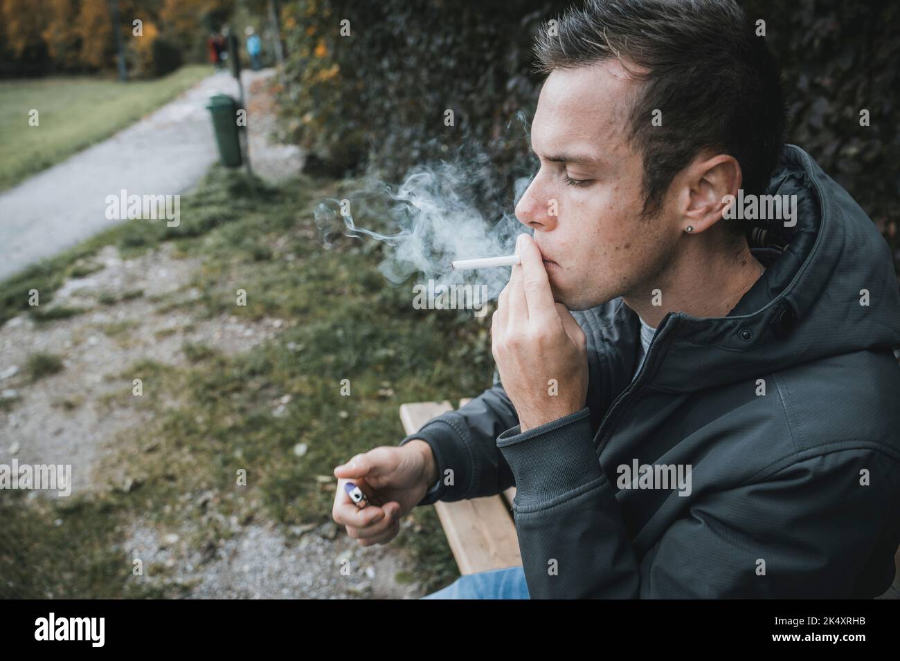 03 de octubre de 2022: Joven sentado en un banco del parque Fumando un cigarrillo. Fumador Enciende Un cigarrillo con encendedor para fumar Foto de stock