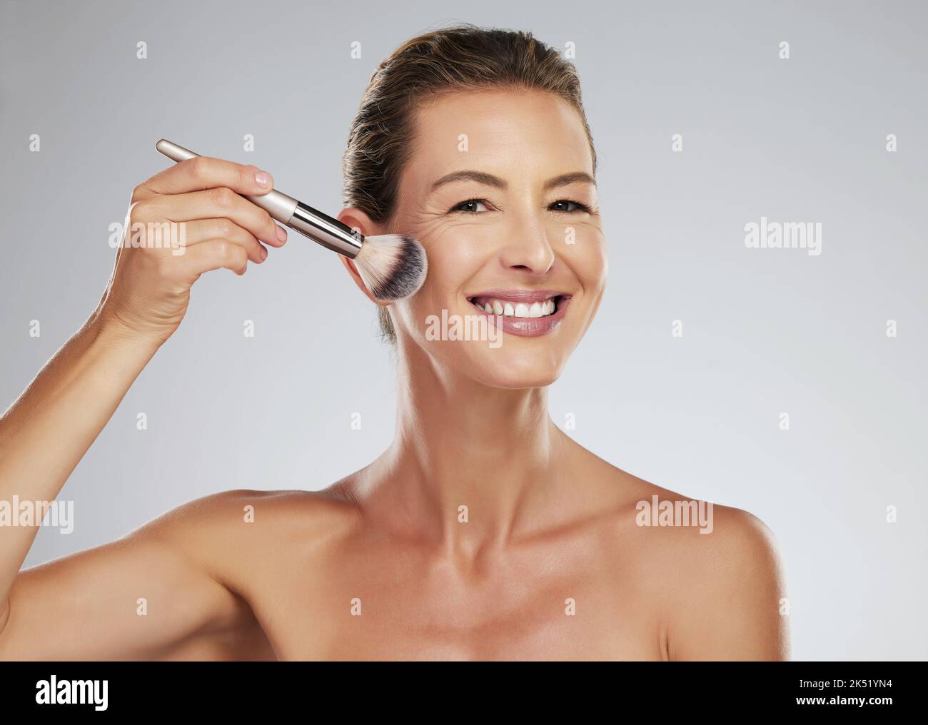 Cara, maquillaje y cepillo con una mujer madura aplicando blusher a su mejilla en el estudio sobre un fondo gris. Belleza, piel y cosméticos con un atractivo Foto de stock