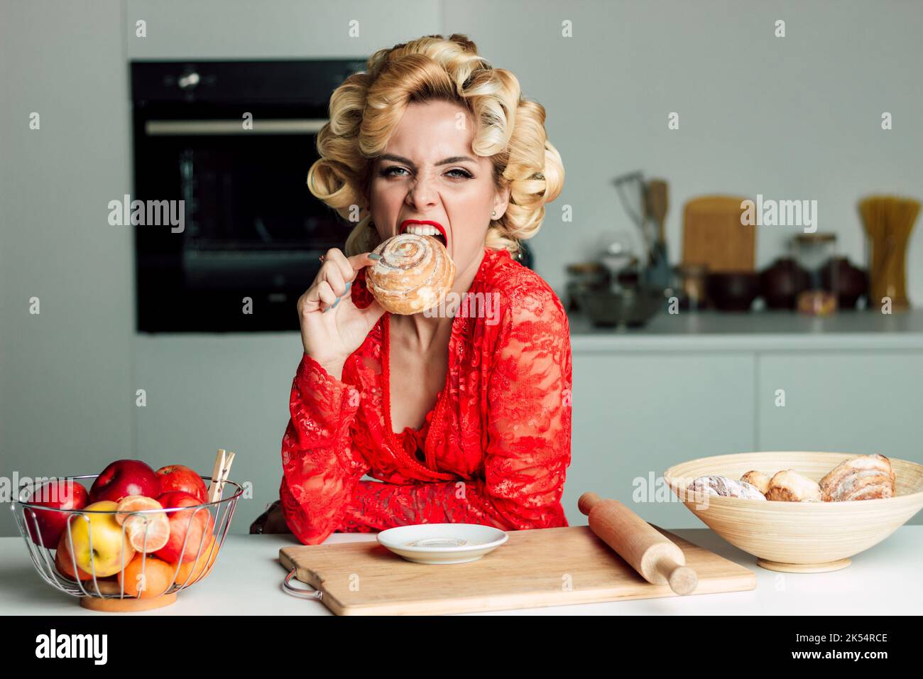 Mujer hermosa en ropa roja muerde un bollo en la cocina en la mesa Foto de stock