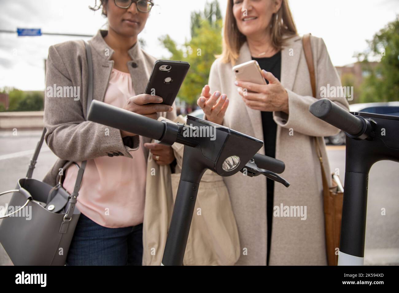Mujeres de negocios con teléfonos inteligentes que alquilan scooters públicos Foto de stock