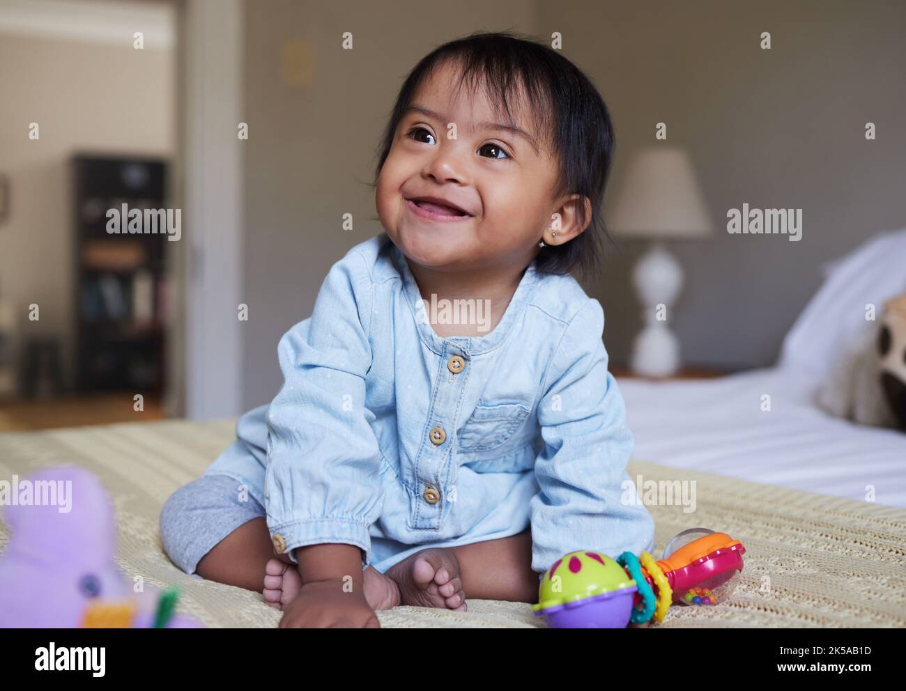 Síndrome de Down, sonrisa y bebé feliz Relájate en la cama divirtiéndose, jugando con juguetes y disfrutando de la felicidad en casa. Discapacidad intelectual, discapacidad mental Foto de stock