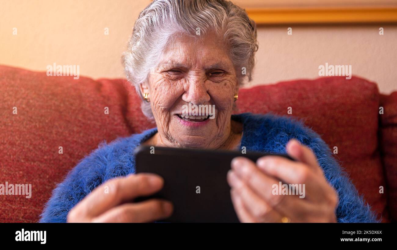 Mujer mayor que usa un teléfono móvil mientras está sentada en el sofá de su casa. La abuela sonriente y feliz sostiene un smartphone. Vieja abuela que opera un teléfono. Concep Foto de stock