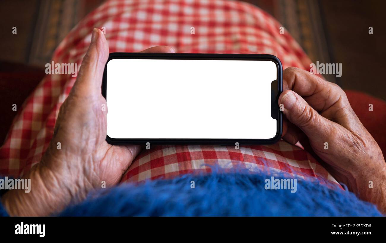 Una anciana en casa usando un teléfono móvil con pantalla blanca. Mockup dispositivo para añadir elementos. Las manos arrugadas de la abuela sostienen un smartphone. Concepto de Foto de stock