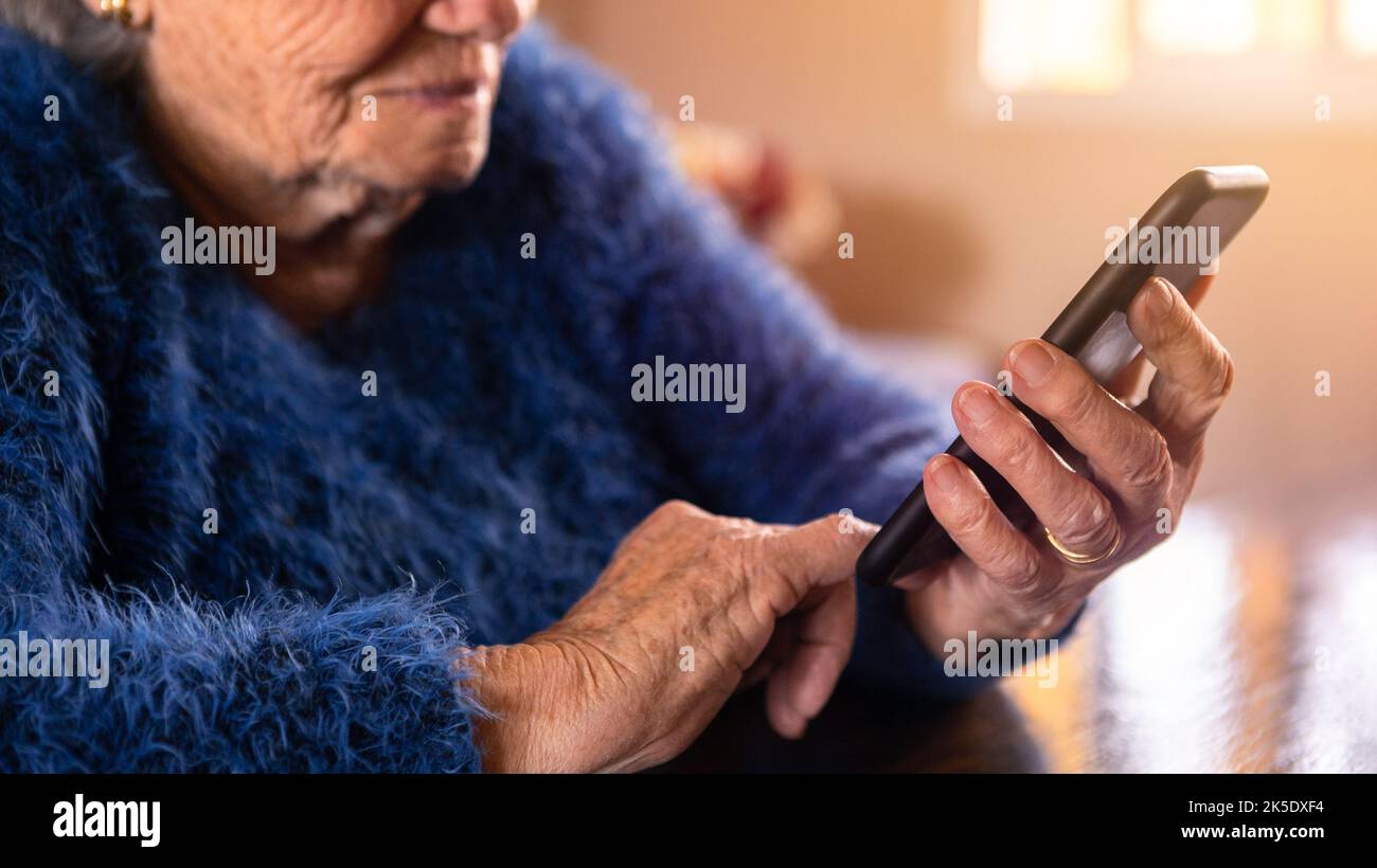 Mujer anciana que utiliza un teléfono móvil mientras está sentada en la casa del salón. La abuela tiene un smartphone. Vieja abuela que opera un teléfono. Concepto de te Foto de stock