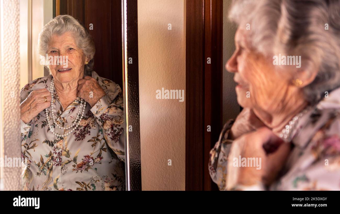 Feliz y alegre abuela caucásica 90s en ropa elegante mirando al espejo. La abuela se siente optimista en la casa de la sala de estar. Concepto de personas mayores. Foto de stock