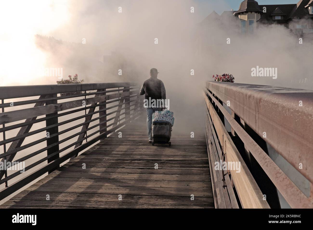 Un hombre camina con equipaje a través de un puente peatonal mientras los gases de las fuentes termales geotérmicas naturales envuelven el paisaje en Pagosa Springs, Colorado, EE.UU. Foto de stock
