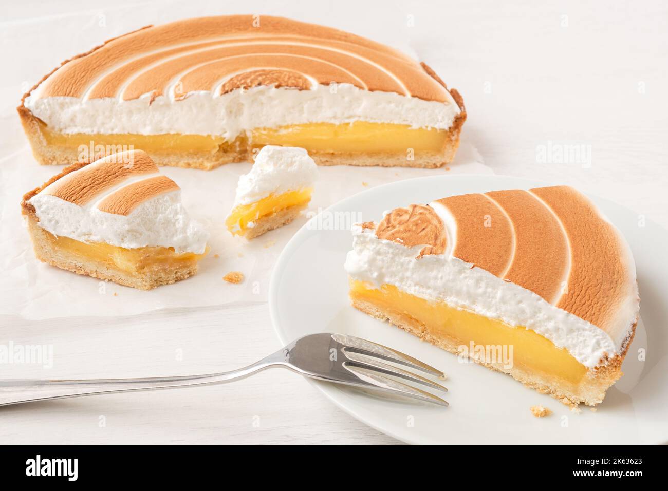 Corte la tarta de limón con merengue encima en papel de hornear junto a una porción de tarta de limón en el plato con un tenedor. Foto de stock
