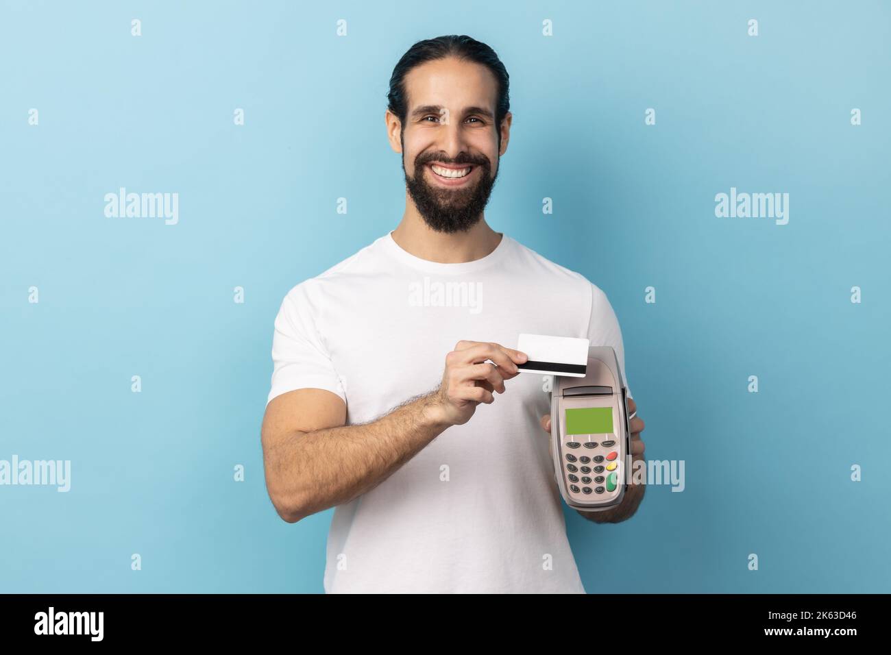 Retrato de hombre sonriente con barba con camiseta blanca sujetando en la mano pos terminal de pago, va a hacer el pago con tarjeta, compras sin efectivo. Estudio de interior aislado sobre fondo azul. Foto de stock