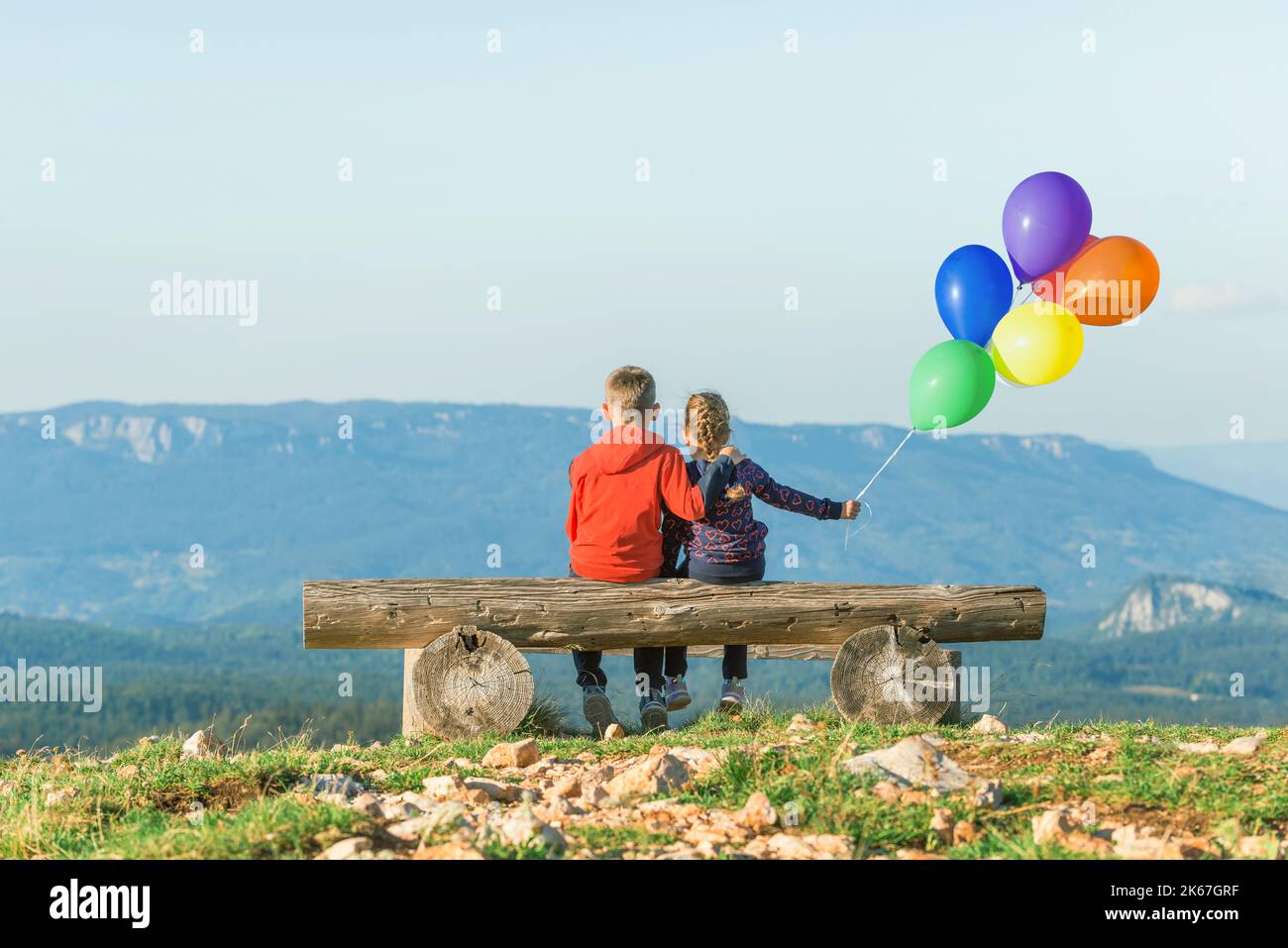 Una niña y un niño están sentados en un banco con globos balanceándose en el viento. Vista desde la parte de atrás. Montaña en el fondo Foto de stock