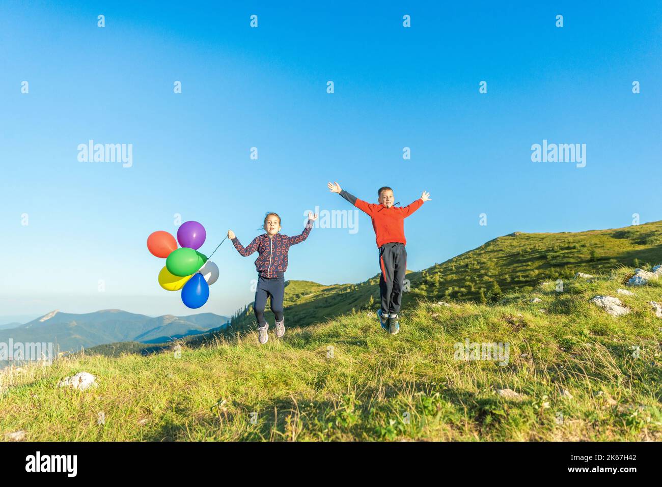 Una niña y un niño saltando con los brazos extendidos con globos en la ladera de la montaña. El concepto de manifestación de la felicidad Foto de stock