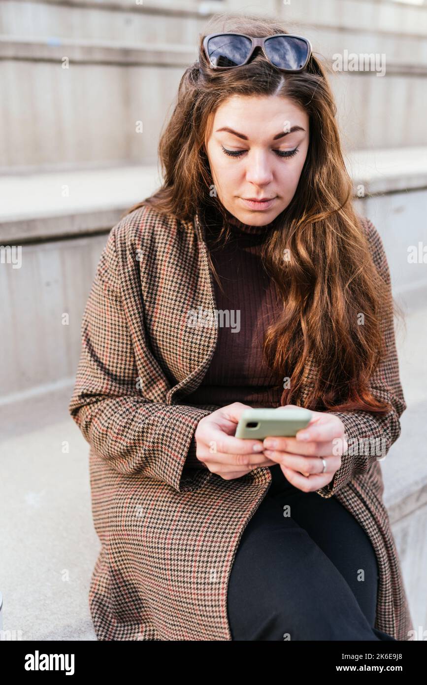 Mujer de smartphone de navegación de pelo largo mientras se sienta en los escalones de la calle de la ciudad por la Fotografía de stock - Alamy