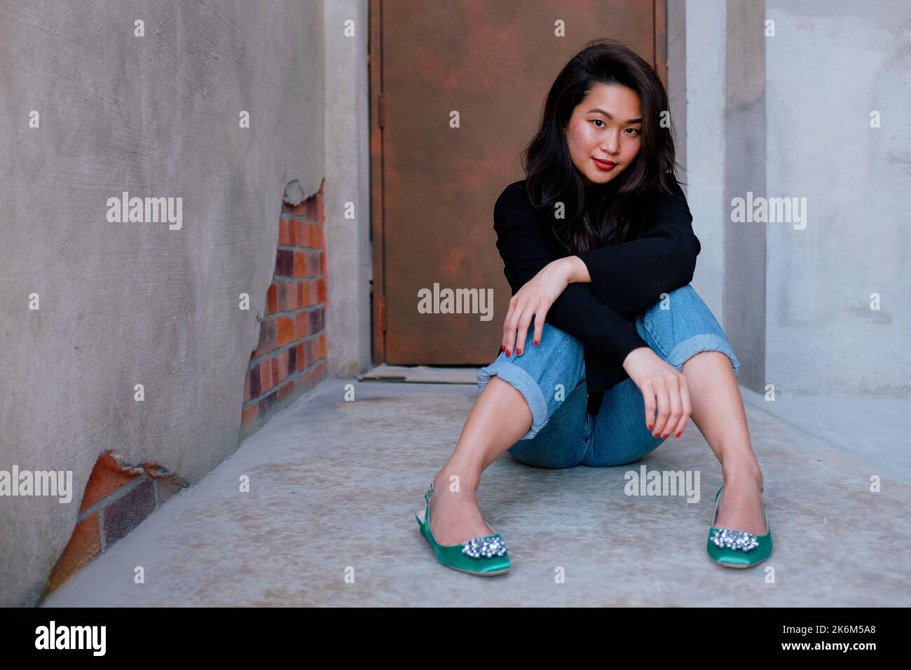Bonita mujer del sudeste asiático que usa un suéter negro, vaqueros y zapatos verdes se sienta en el suelo fuera de una puerta del escenario. La mujer mira a la cámara. Foto de stock