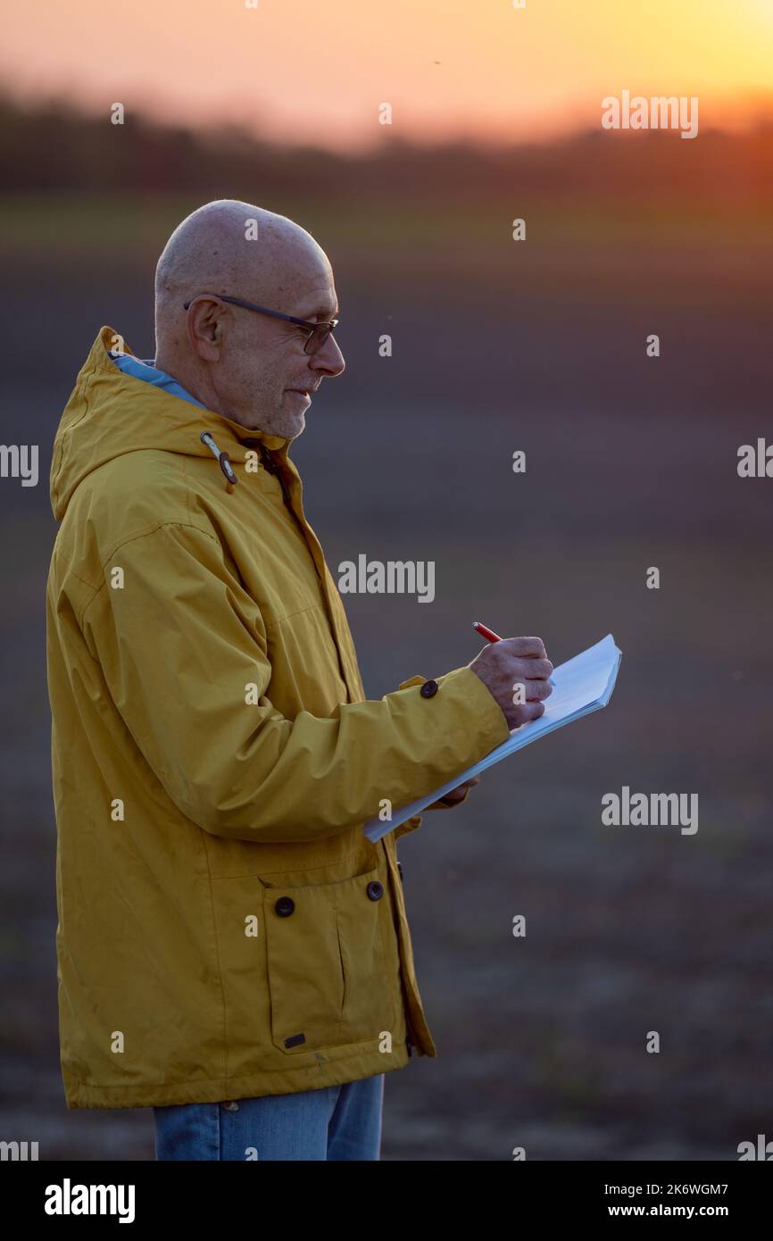 Granjero senior parado en el campo al anochecer y escribiendo notas a finales de septiembre Foto de stock