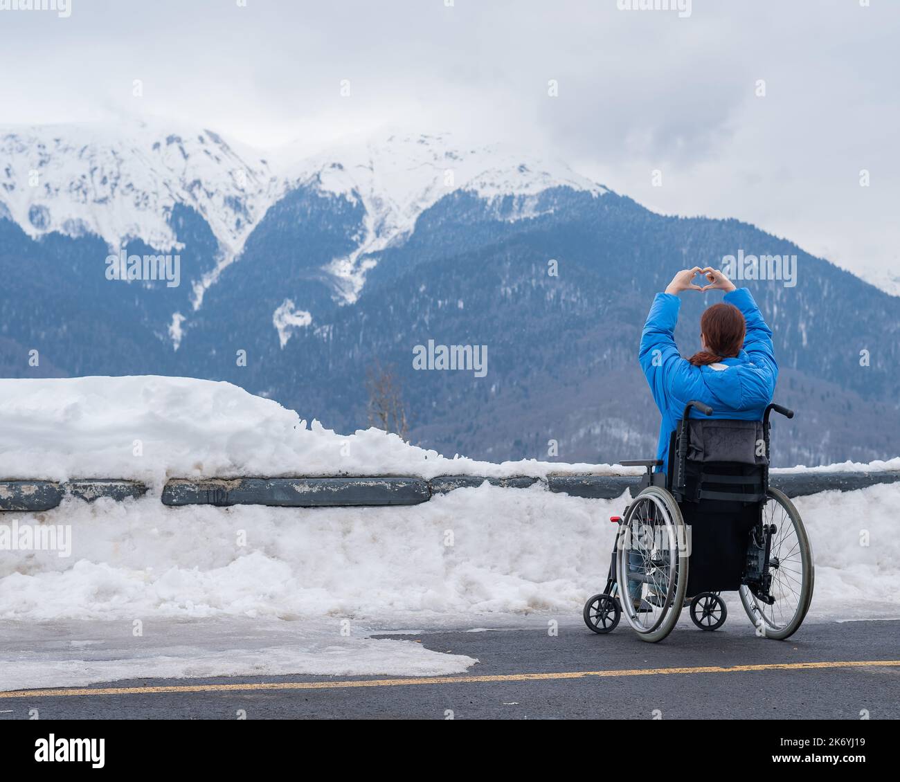 En invierno, una mujer en silla de ruedas sostiene las manos en forma de corazón en las montañas. Foto de stock