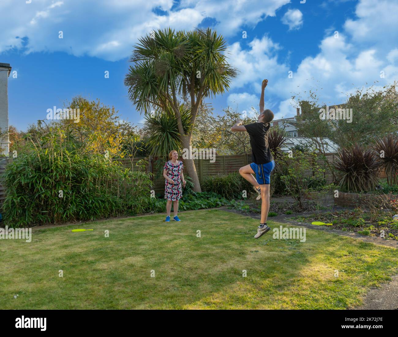 Adolescente con equipo deportivo que salta a la altura para atrapar una pelota de tenis jugando con su madre en el jardín de casa Foto de stock