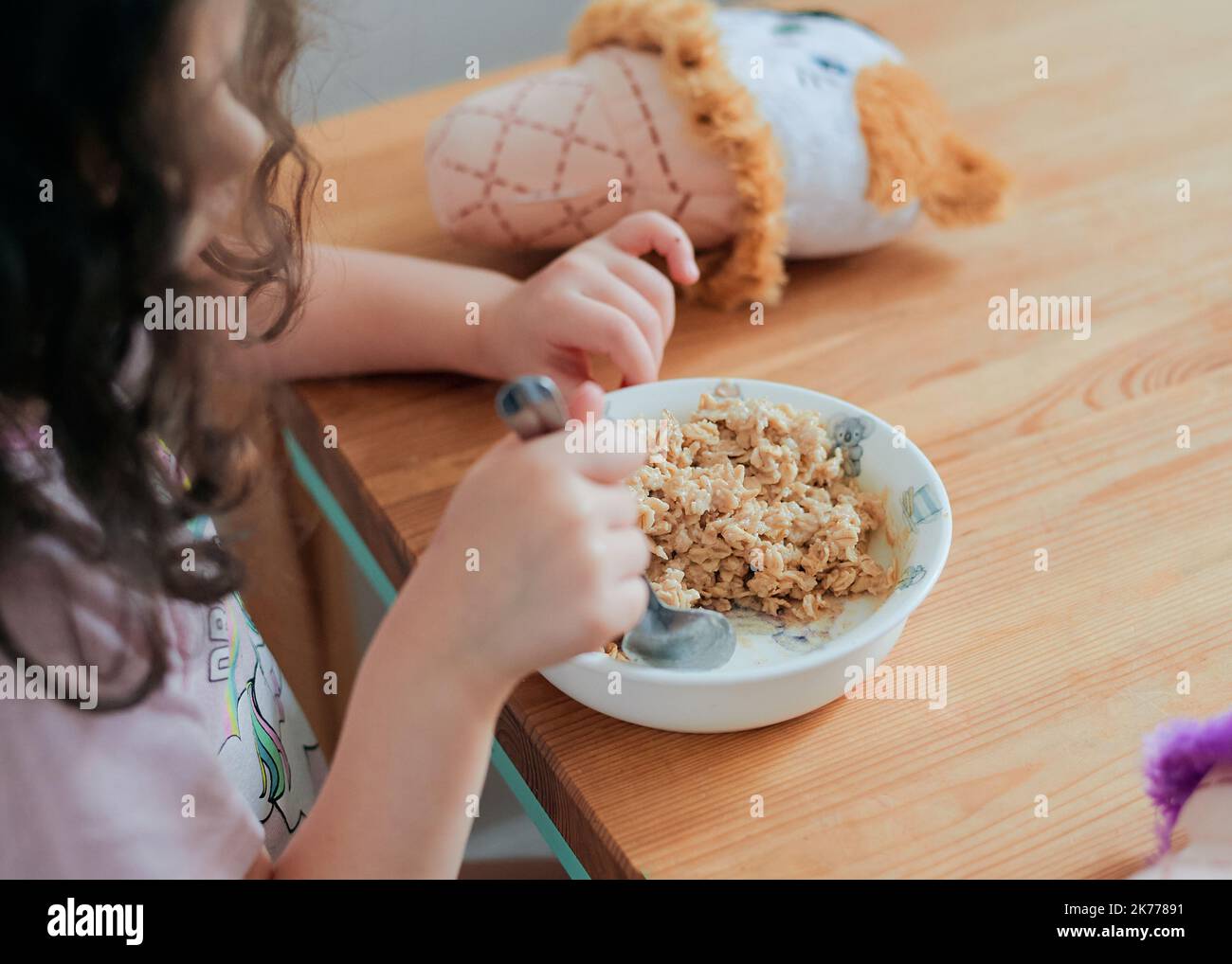 una chica toma un desayuno de avena en la mesa de la cocina rodeada de juguetes Foto de stock