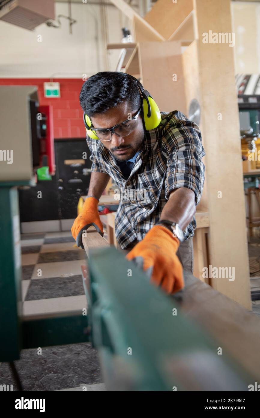 Hombre joven que lleva protectores auditivos aserrando madera en el espacio de fabricación Foto de stock