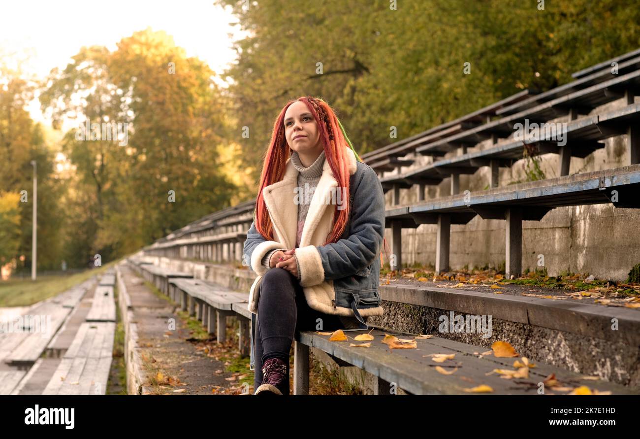 Mujer joven positiva con los dreadlocks mirando lejos, sonriendo, sentado en viejo banco destartalado de la calle bleacher en otoño de oro Foto de stock