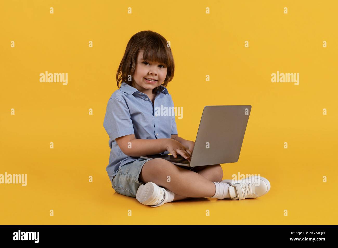 Adorable niño escribiendo en el portátil, disfrutando de la educación en línea, sentado en el suelo y sonriendo a la cámara, espacio libre Foto de stock