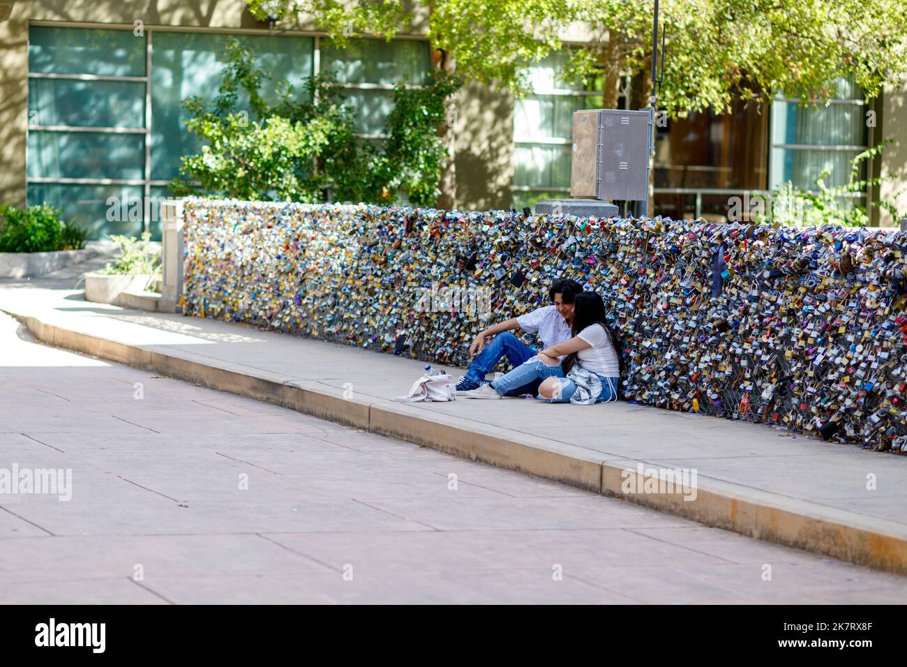 Una pareja se sienta una al lado de la otra en una acera con cientos de cerraduras de amor detrás de ellos a lo largo del River Walk en el centro de San Antonio, Texas. Foto de stock