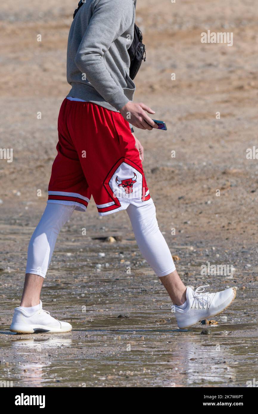 Un hombre de vacaciones con pantalones cortos de baloncesto Chicago Bulls Red Just Don sobre leggings blancos y zapatillas caminando por la playa Fistral en Newquay en Cornw Foto de stock