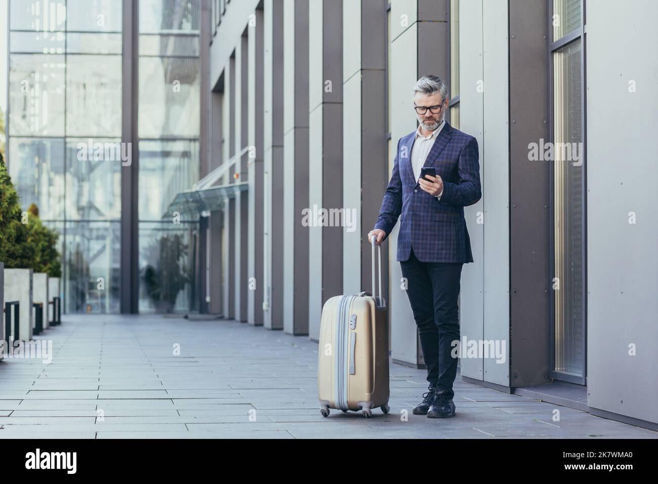 Viaje de negocios, hombre de negocios mayor de pelo gris fuera del hotel utilizando el teléfono, jefe maduro con maleta grande, inversor en traje de negocios y gafas. Foto de stock