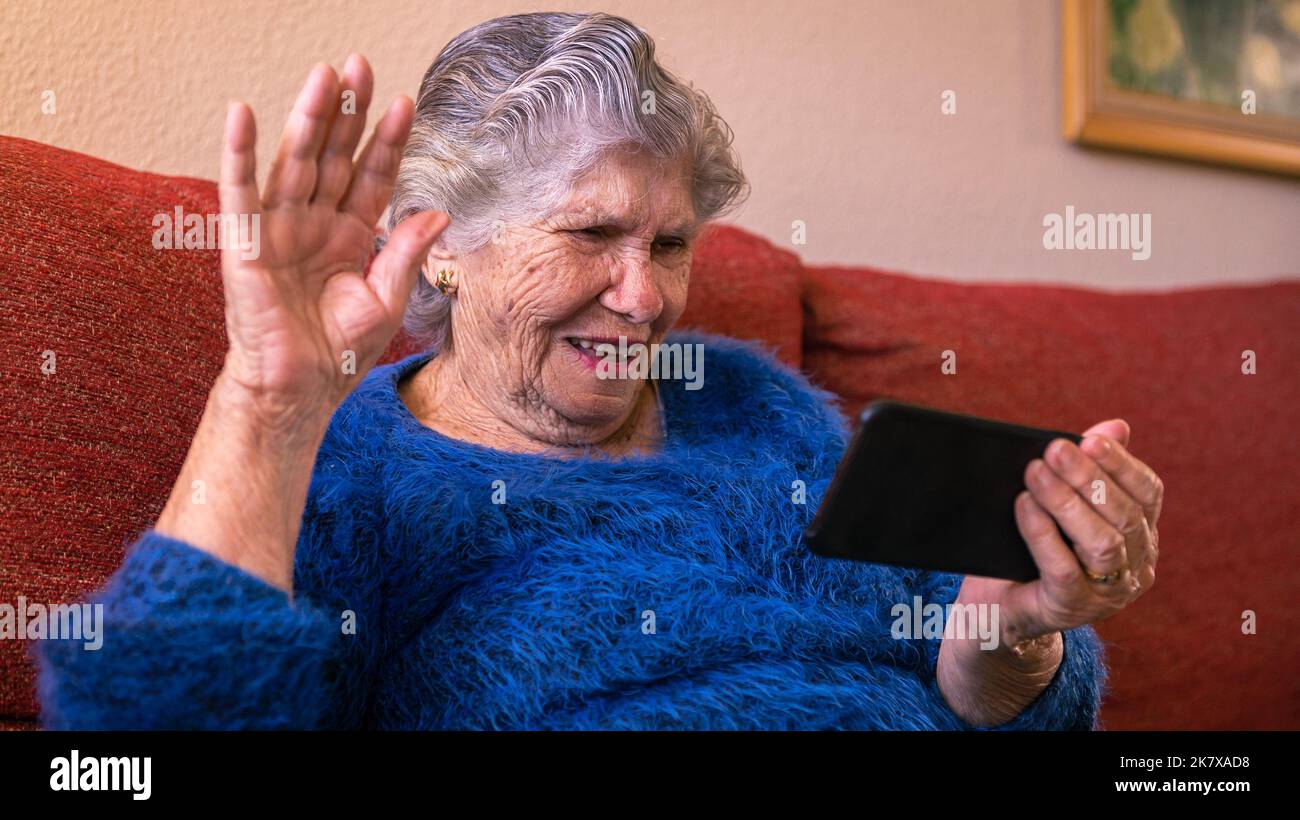 Feliz sonrisa anciana caído de la mano en el saludo mientras utiliza un smartphone para hablar. Abuela caucásica hablando por teléfono móvil en la casa de la sala de estar. G Foto de stock
