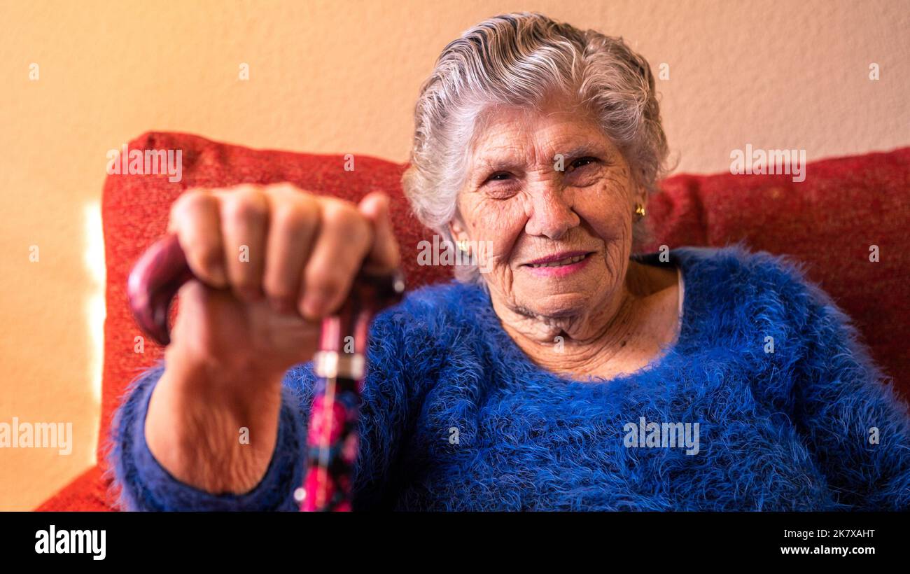 Vieja mujer caucásica de 90s años sosteniendo un bastón y sentirse feliz y sonriente en el sofá de la sala de estar. La mujer alquiladora o abuela se relajan en el sofá de la casa. Foto de stock