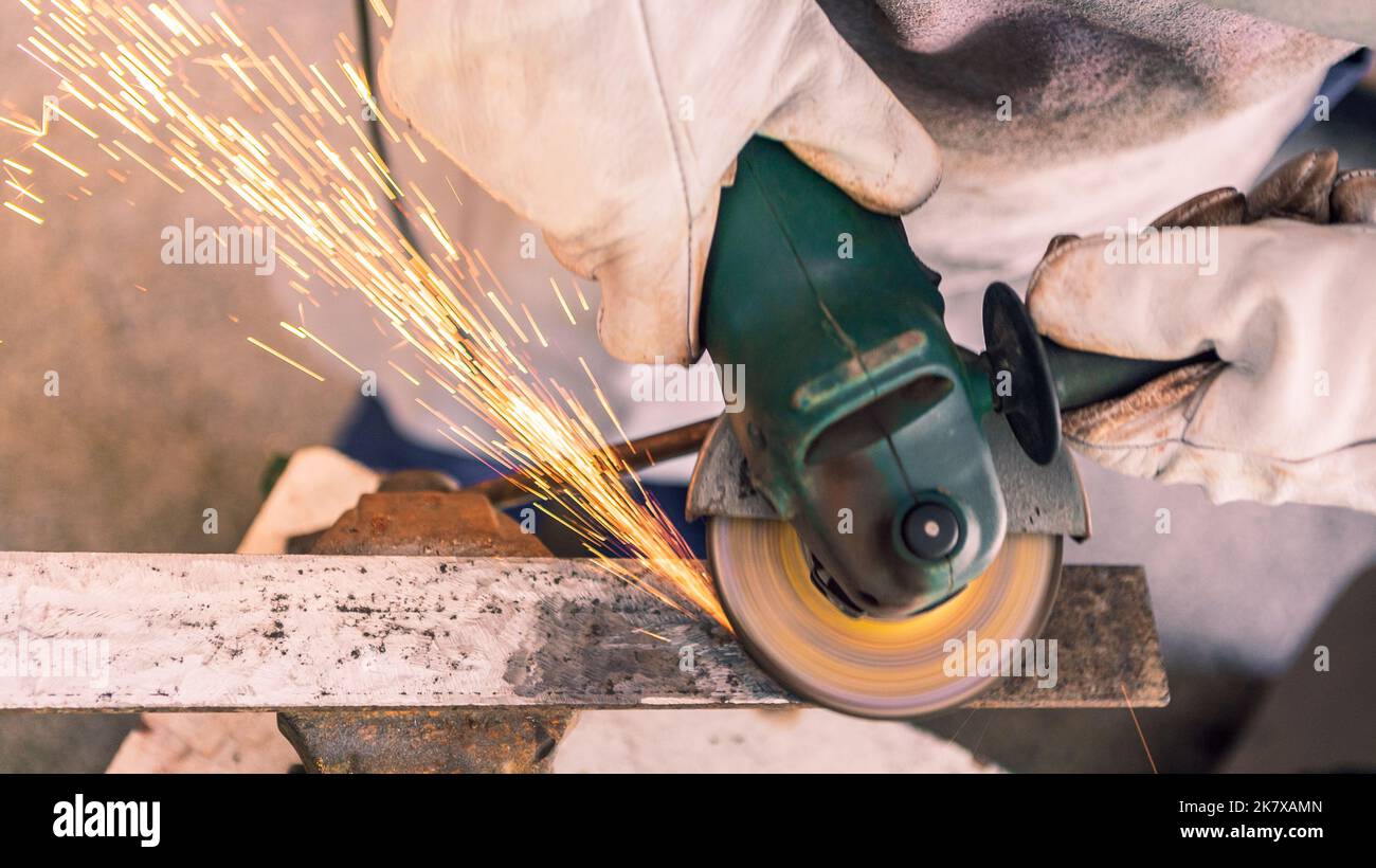 Procesamiento de metal con un hombre que utiliza una amoladora angular para limpiar la costura de acero. Cerrajero en ropa especial y guantes trabaja en producción. Chispas en el MET Foto de stock