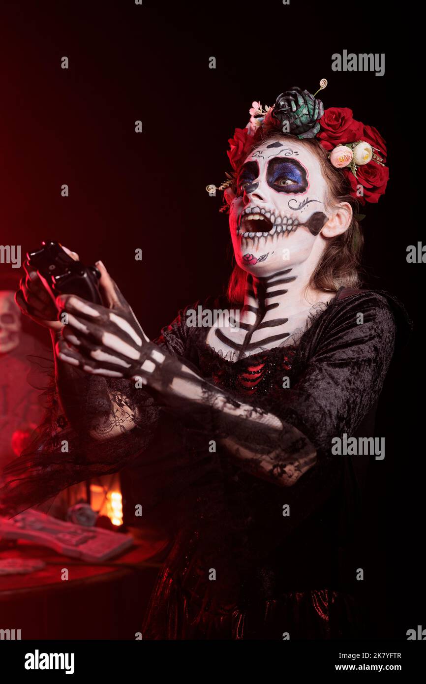 Goddess of Death Jugando videojuegos online con joystick en la consola,  usando maquillaje de calavera y traje tradicional de santa muerte. Mujer  disfrutando de la competencia de juegos el día de los