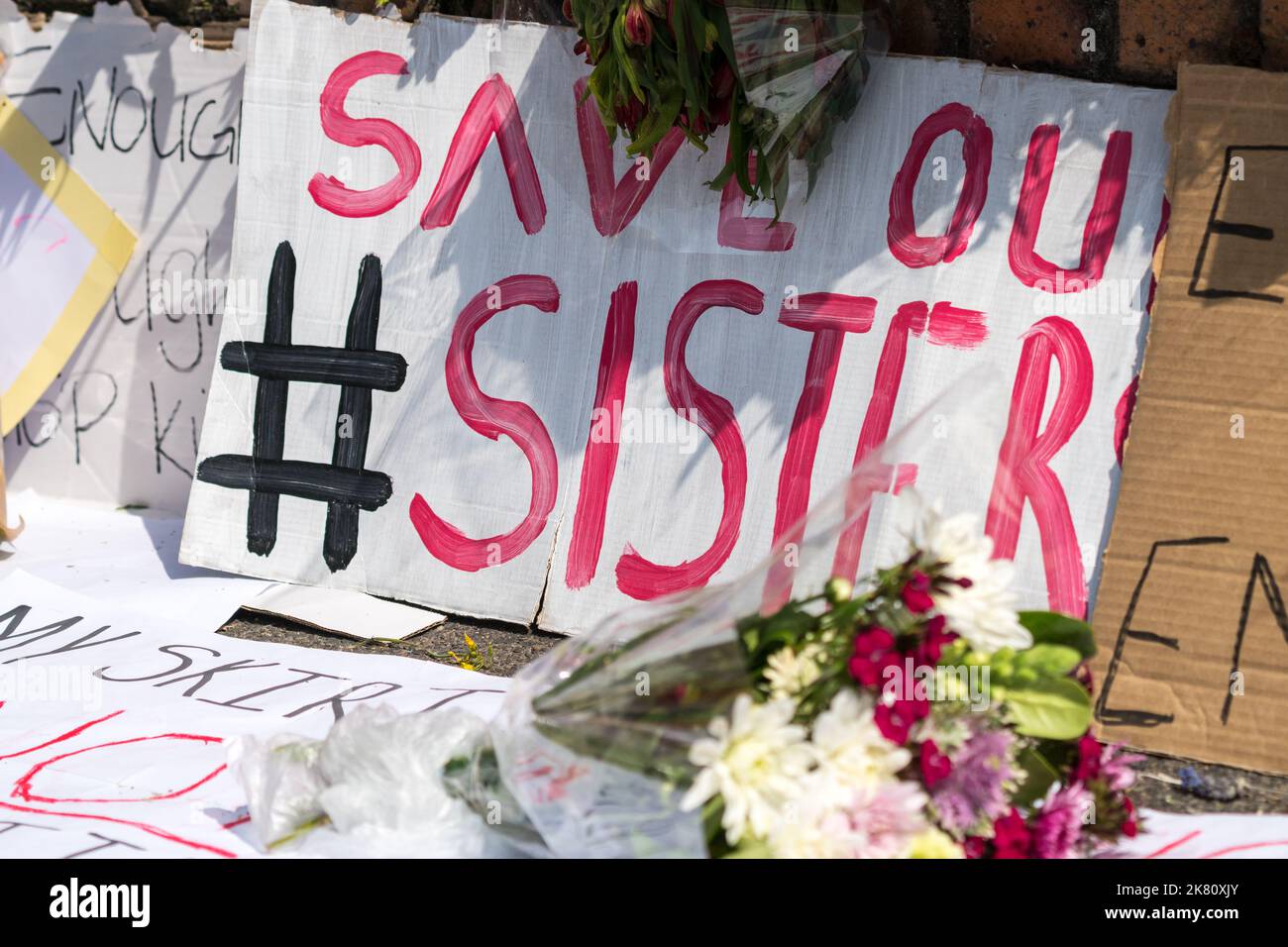 Hashtag salvar a nuestras hermanas, letrero pintado a mano rodeado de mensajes de simpatía y flores en un pavimento o acera en Sudáfrica Foto de stock